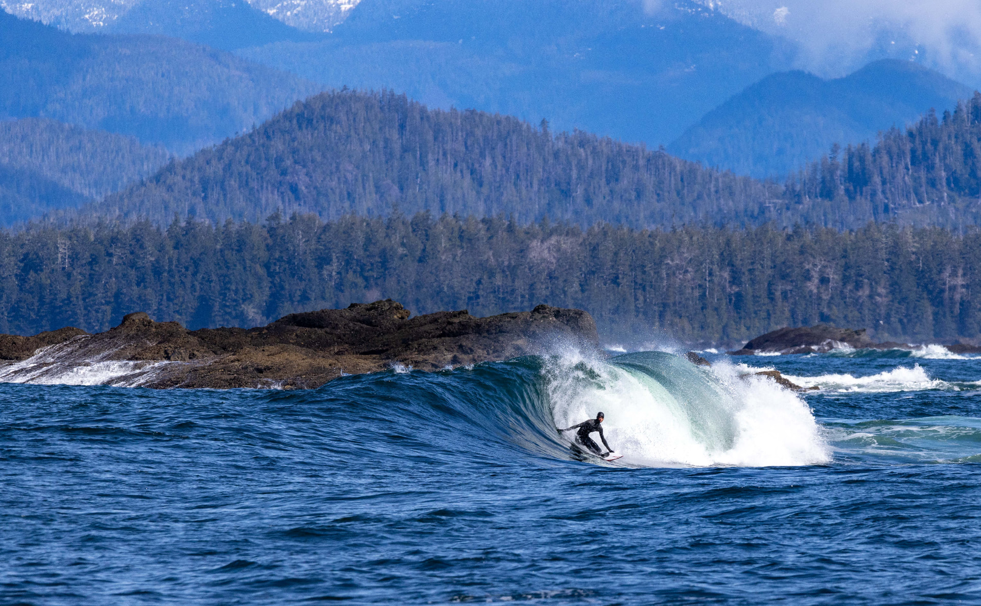 A surfer riding a clean wave off a rocky coastline below forested mountains