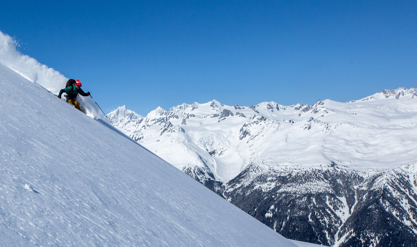 A skier carving deep powder on a steep face above snow-covered peaks