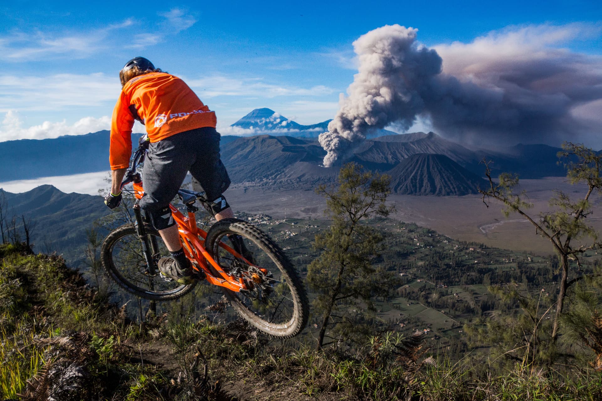 A mountain biker airborne on a ridgeline above an erupting volcano